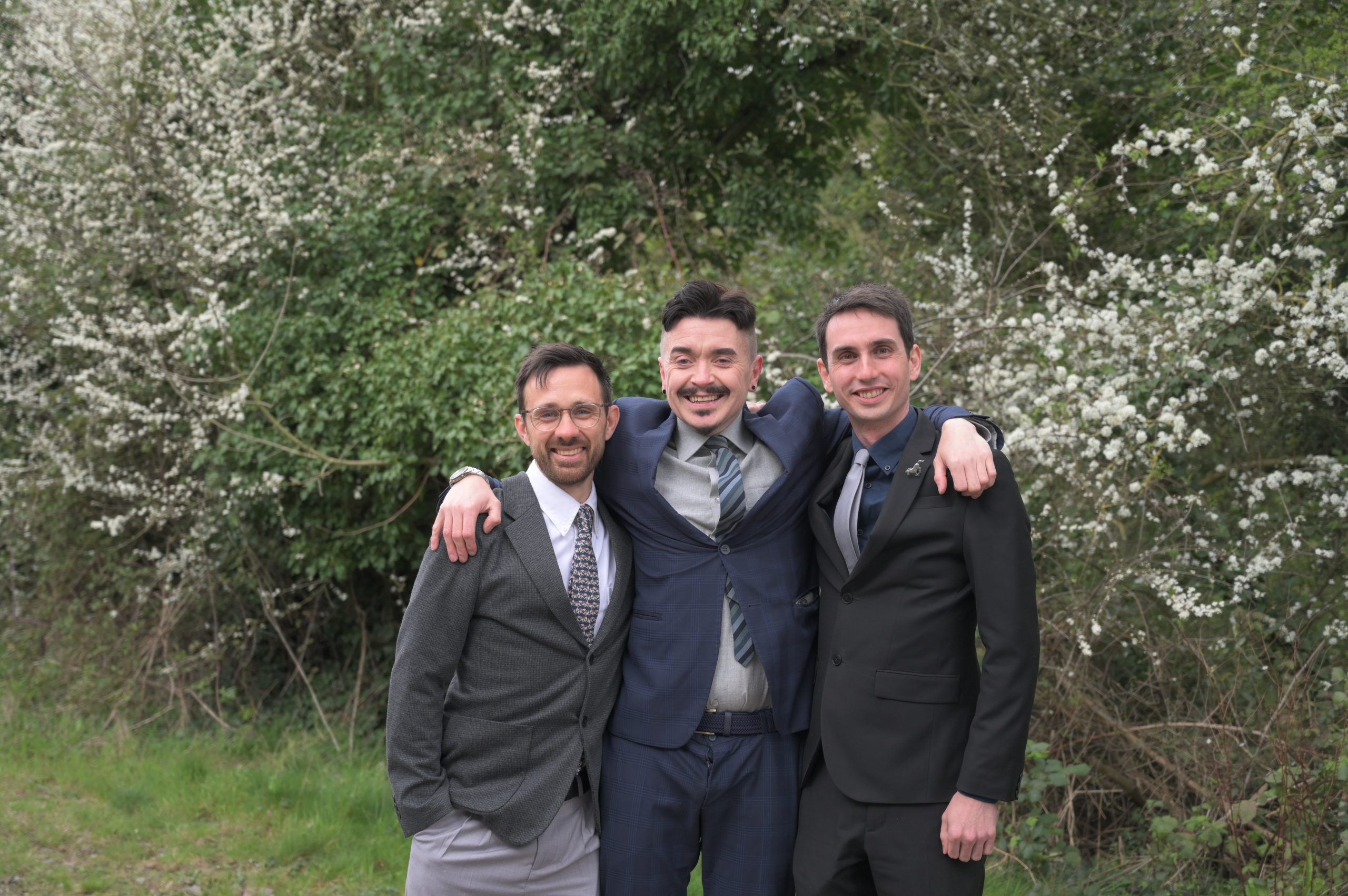 Three men in suits smile with arms around each other in front of blooming trees. The mood is joyful and the setting is a lush, green garden.