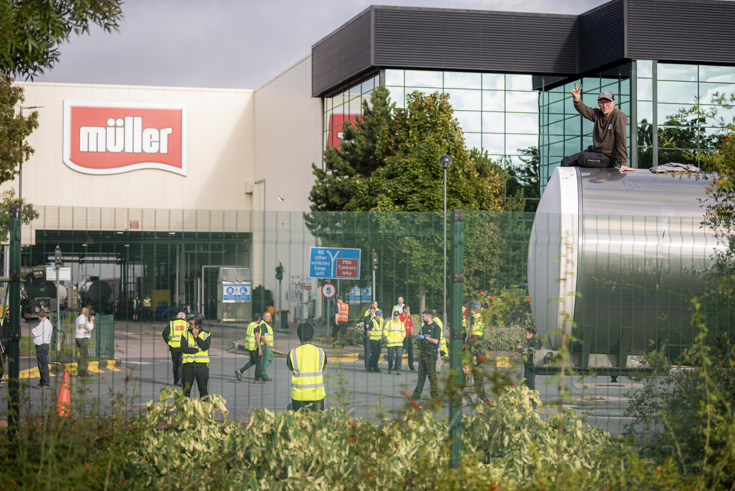 A man sits atop a milk truck making a peace sign with his fingers at a Muller factory while police and security officers mill around the area