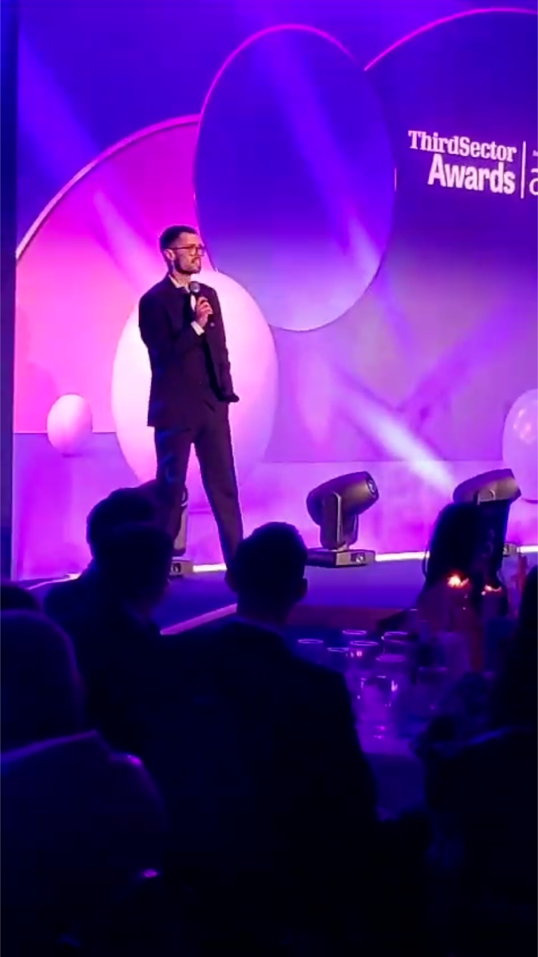 A smartly dressed man stands on stage, microphone in hand, at the ThirdSector Awards ceremony